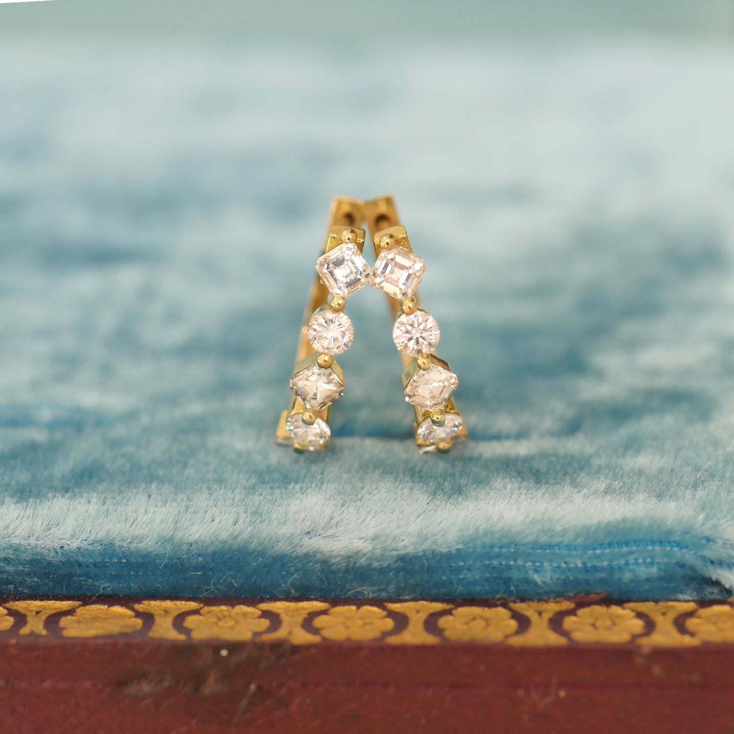 A pair of vintage diamond hoop earrings with six asscher cut diamonds and four round diamonds set in 18k yellow gold, displayed on a blue velvet cushion.